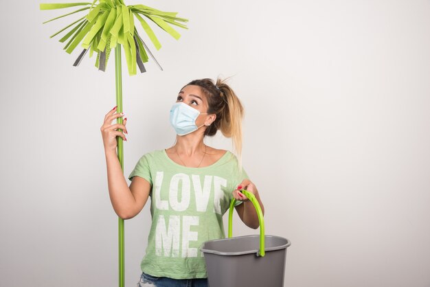 Young female with facemask looking at mop on white wall. 