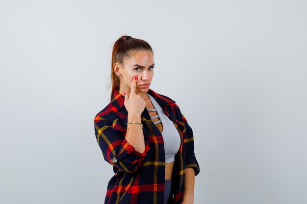 Young female with finger on cheek in crop top, checkered shirt, pants and looking pretty , front view.