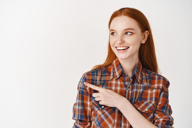 Young female with ginger hair pointing finger left, looking at copy space and smiling, standing over white wall