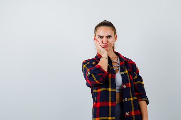 Free photo young female with hand on cheek in crop top, checkered shirt and looking wistful. front view.