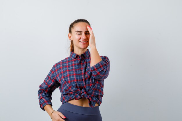 Young female with hand on forehead in checkered shirt, pants and looking happy , front view.