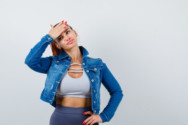 Young female with hand on forehead in crop top, jacket, pants and looking forgetful , front view.