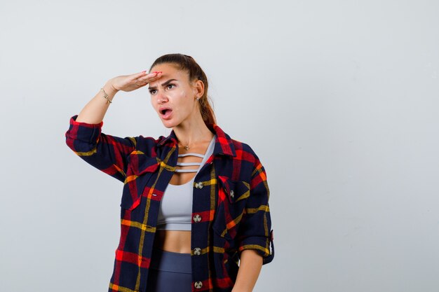 Young female with hand on head to see clearly in crop top, checkered shirt, pants and looking puzzled , front view.