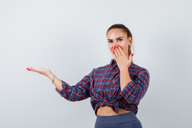 Young female with hand on mouth while showing something in checkered shirt, pants and looking amazed. front view.