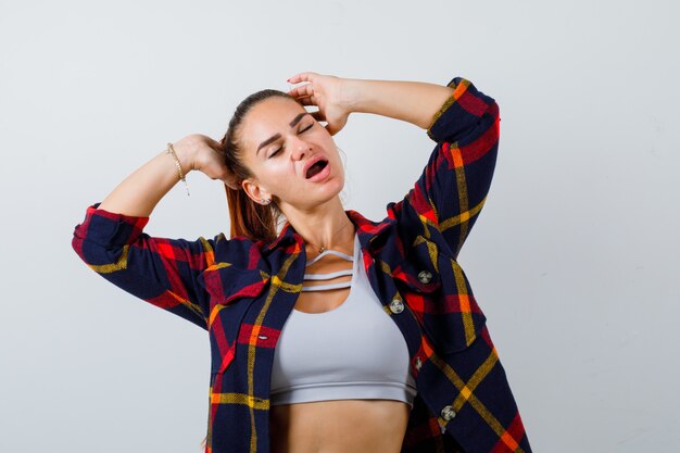 Foto gratuita giovane donna con le mani sulla testa in crop top, camicia a scacchi e dall'aspetto rilassato. vista frontale.