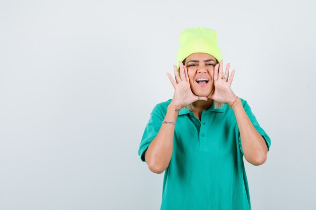 Young female with hands near mouth in polo t-shirt, beanie and looking merry , front view.