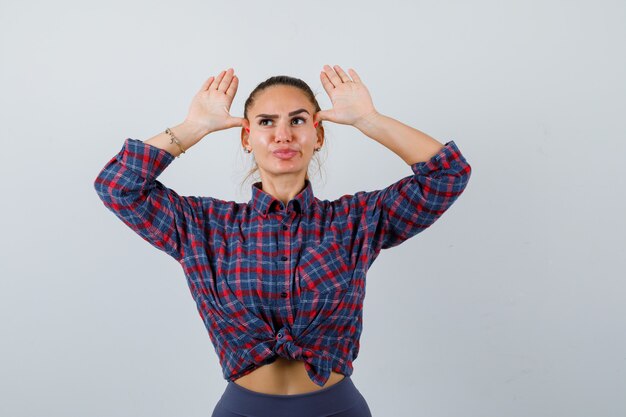 Бесплатное фото Молодая женщина с руками над головой как уши в клетчатой рубашке, штанах и выглядит смешно. передний план.