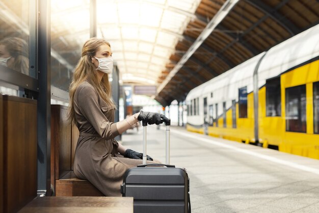 Молодая женщина с чемоданом в маске и перчатках и ждет на вокзале