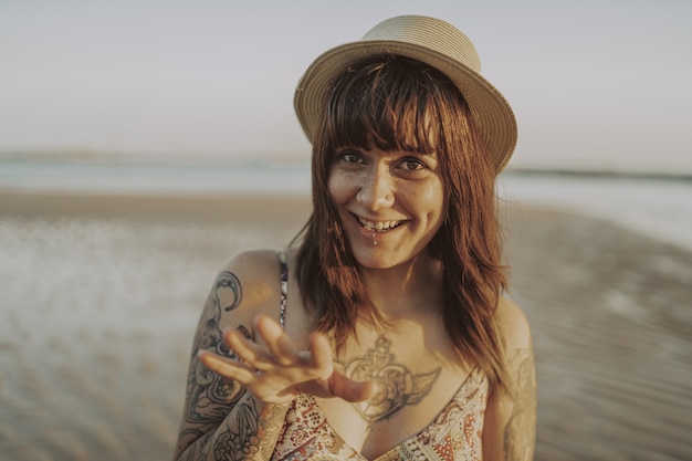 Young female with tattoos wearing a dress and straw hat on blurred ocean