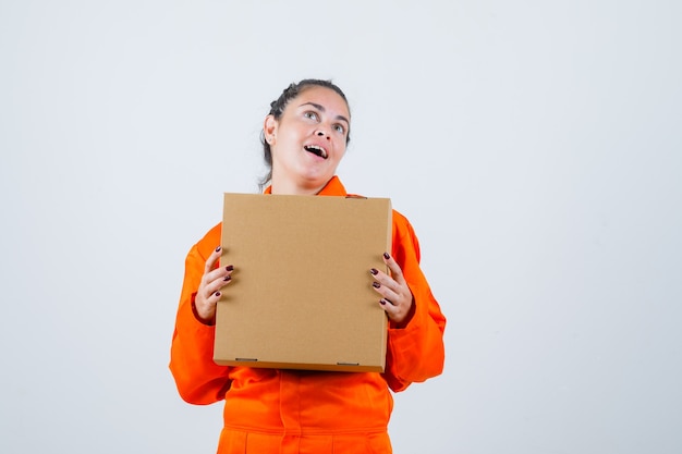 Young female in worker uniform holding box while making a wish and looking ready , front view.