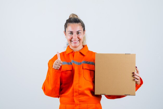 労働者の制服を着た若い女性が箱を持って陽気に見ながら親指を立てて、正面図。