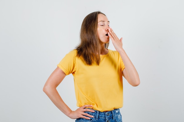 Young female yawning with hand on waist in t-shirt, shorts and looking sleepy. front view.