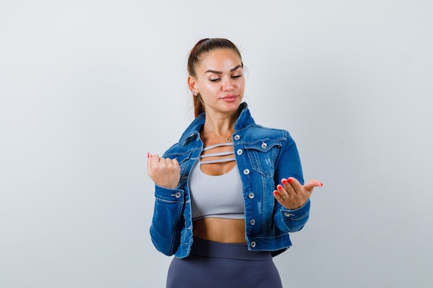 Young fit female looking at spreading palm while showing clenched fist in top, denim jacket and looking confused , front view.