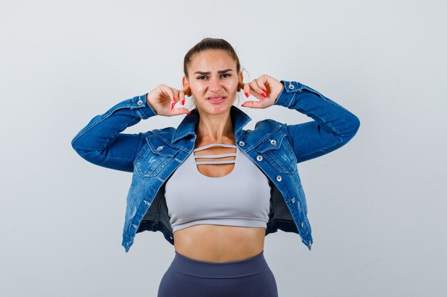 Young fit female plugging ears with fingers in top, denim jacket and looking annoyed , front view.