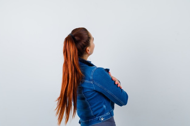 Young fit female in top, denim jacket hugging herself and looking pensive , back view.