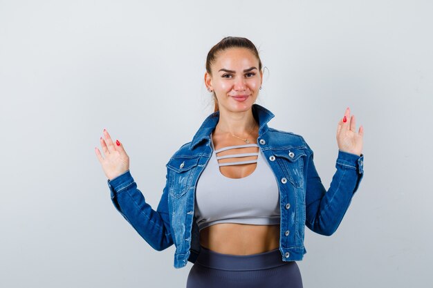 Young fit female in top, denim jacket showing surrender gesture and looking merry , front view.