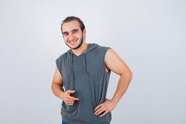 Young fit male posing with hand on waist in sleeveless hoodie  and looking cheerful. front view.