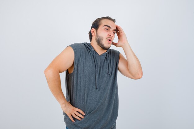 Young fit male posing with hand on waist while keeping hand on face in sleeveless hoodie  and looking wondered. front view.