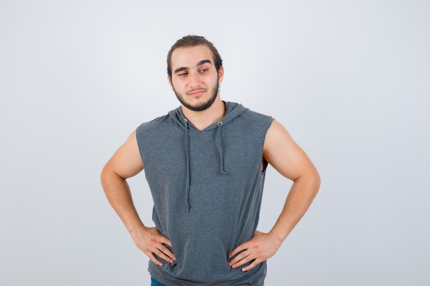 Young fit male posing with hands on waist in sleeveless hoodie  and looking blissful. front view.