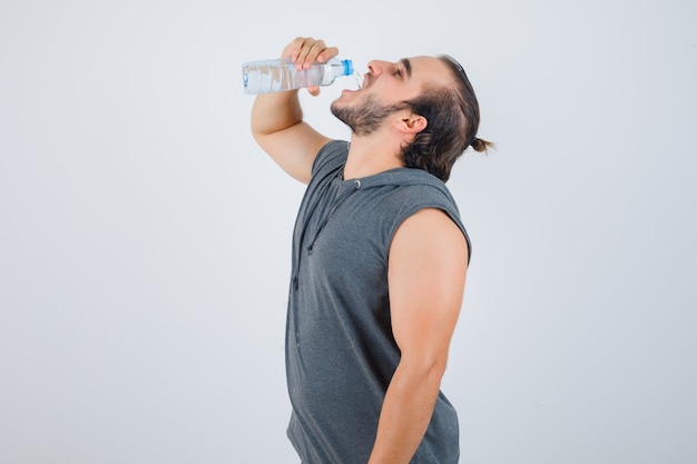 Young fit male in sleeveless hoodie  drinking water and looking merry , front view.