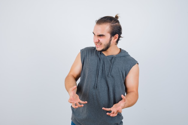 Young fit male in sleeveless hoodie  keeping hands in aggressive manner and looking annoyed , front view.