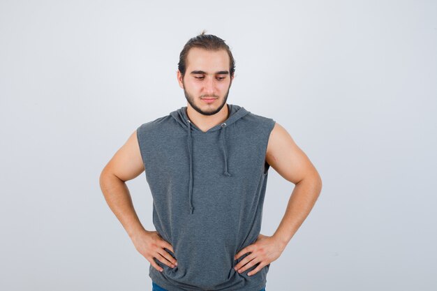 Young fit male in sleeveless hoodie  posing with hands on waist and looking pensive , front view.