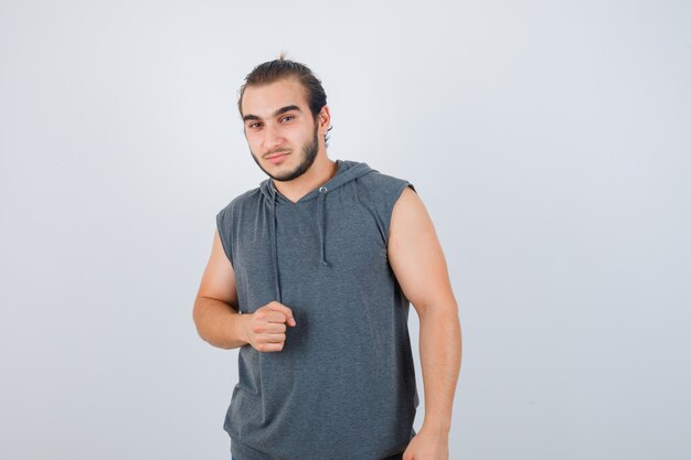 Young fit male in sleeveless hoodie  showing clenched fist and looking confident , front view.