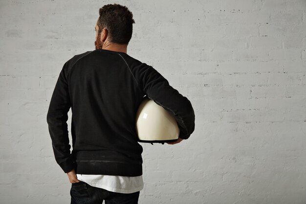 Free photo young fit motorcyclist in black sweatshirt and jeans holding a white helmet on his side with painted brick walls