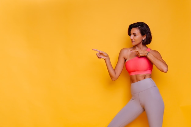 Foto gratuita giovane donna in forma abbastanza forte vestita con abiti sportivi, top alla moda e leggings, posa contro il muro giallo