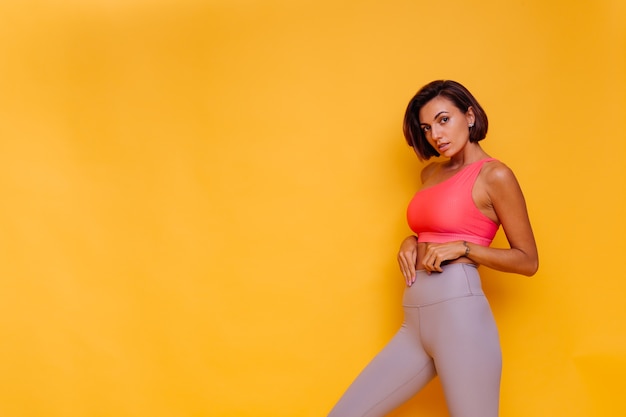 Young fit pretty strong woman dressed in sport clothes, stylish top and leggings, poses against yellow wall