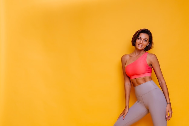 Free photo young fit pretty strong woman dressed in sport clothes, stylish top and leggings, poses against yellow wall