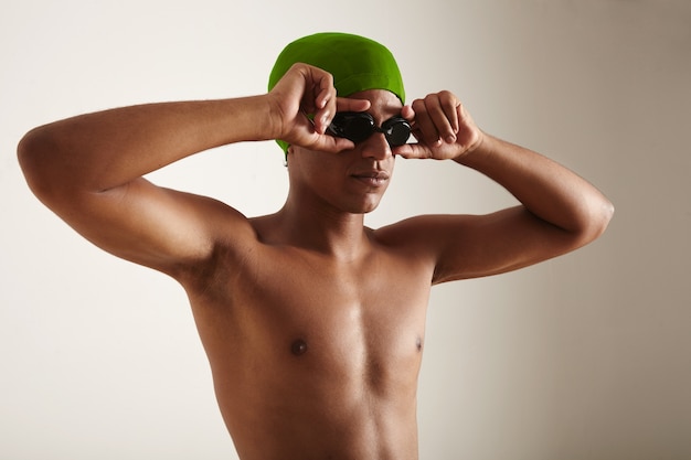 Young fit relaxed black swimmer in green cap putting on black goggles on white