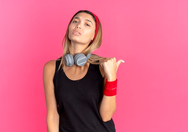 Free photo young fitness girl in black sportswear and red headband eith serious face pointing back over pink