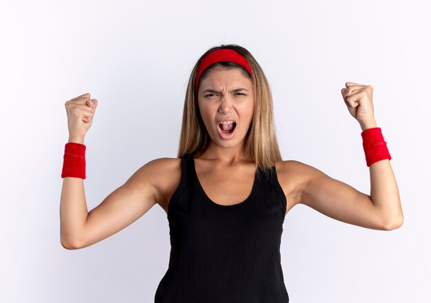 Young fitness girl in black sportswear and red headband going wild clenching and raising fists shouting with angry face standing over white wall