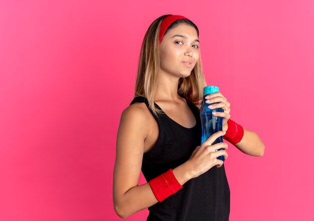 Молодая фитнес-девушка в черной спортивной одежде и красной повязке на голове держит бутылку воды, уверенно улыбаясь над розовым