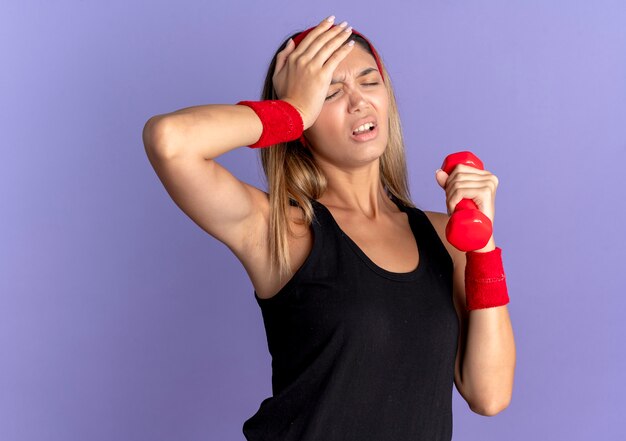 Young fitness girl in black sportswear and red headband holding dumbbell looking tired and exhausted with hnad on head over blue