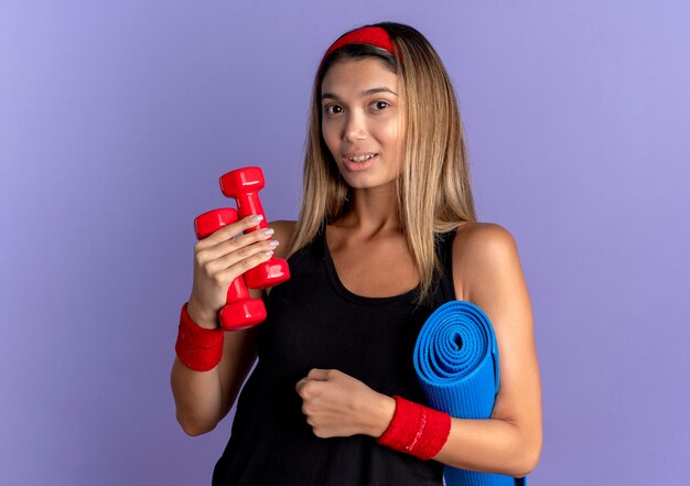 Young fitness girl in black sportswear and red headband holding dumbbells and yoga mat looking at camera smiling confident standing over blue wall