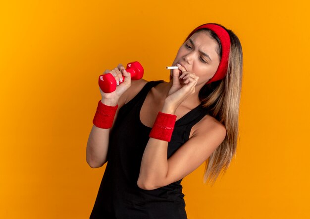 Young fitness girl in black sportswear and red headband holding dummbbell and smoking a cigarette bad habit concept standing over orange wall