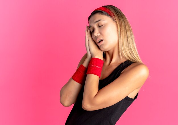Young fitness girl in black sportswear and red headband holding palms making sleep gesture leaning head on palms over pink