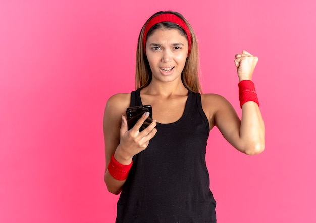 Young fitness girl in black sportswear and red headband holding smartphone clenching fist happy and excited standing over pink wall