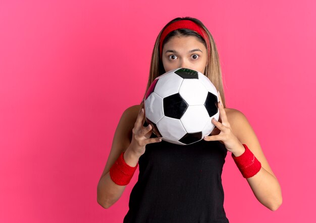 Young fitness girl in black sportswear and red headband holding soccer ball looking surprised hiding her face standing over pink wall