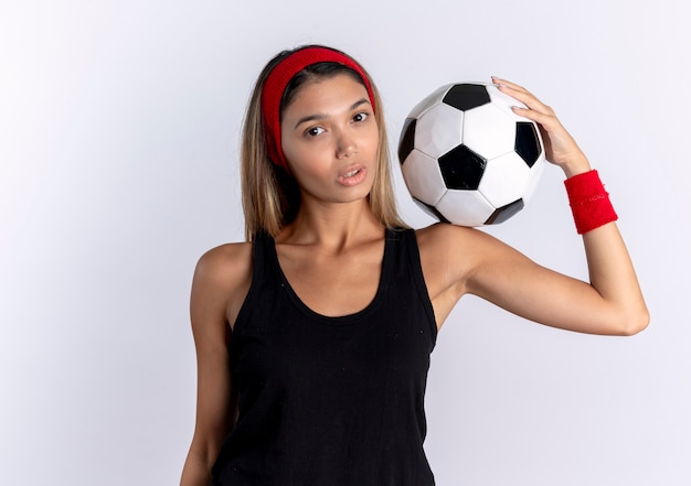 Young fitness girl in black sportswear and red headband holding soccer ball on shoulder  with serious face standing over white wall