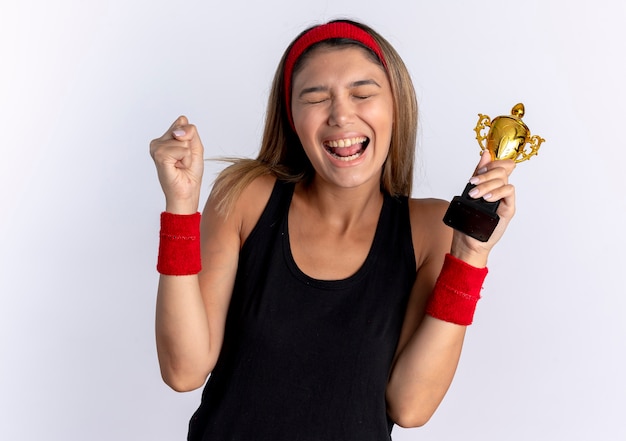 Young fitness girl in black sportswear and red headband holding trophy clenching fist happy and excited standing over white wall