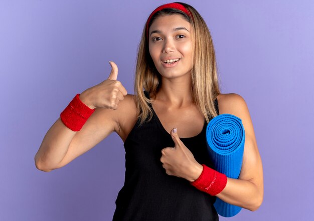 Молодая фитнес-девушка в черной спортивной одежде и красной повязке на голову, держащая коврик для йоги, глядя в камеру, улыбаясь, показывает палец вверх, стоя над синей стеной