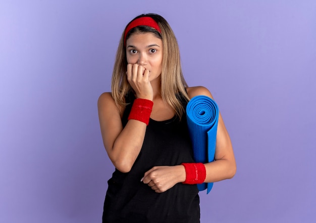 Young fitness girl in black sportswear and red headband holding yoga mat looking stressed and nervous biting nails standing over blue wall