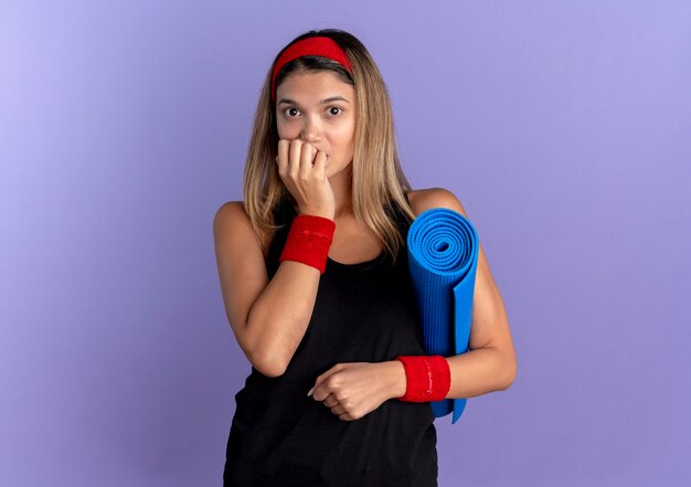 Молодая фитнес-девушка в черной спортивной одежде и красной повязке на голову, держащая коврик для йоги, выглядит напряженной и нервной, кусая ногти, стоя над синей стеной