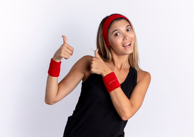 Young fitness girl in black sportswear and red headband lookign  smiling showing thumbs up standing over white wall