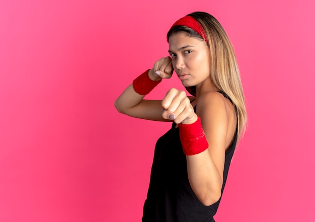 Young fitness girl in black sportswear and red headband looking confident clenching fists with serious face standing over pink wall