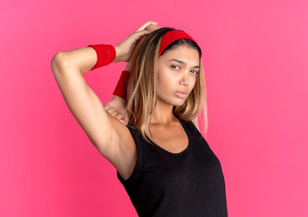 Young fitness girl in black sportswear and red headband looking confident stretching herself over pink