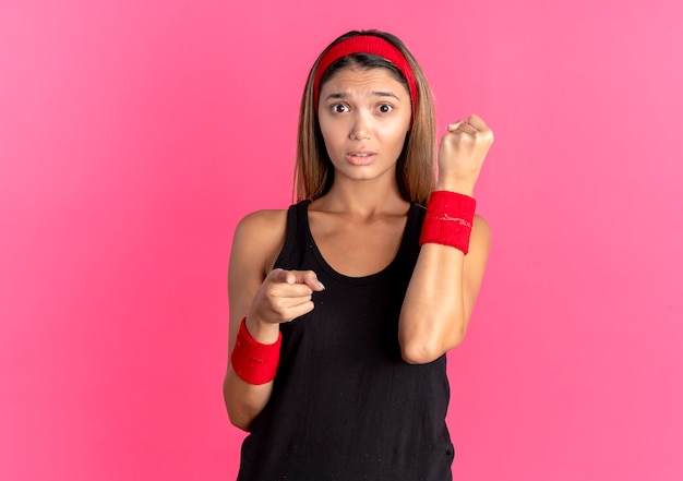 ピンクの上のカメラで指で指している混乱した握りこぶしを探している黒いスポーツウェアと赤いヘッドバンドの若いフィットネスの女の子
